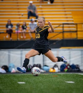 Youth soccer player in black uniform kicks a soccer ball on the field, with spectators in the background.