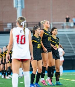 Three female soccer players in black uniforms line up on the field, focused and ready for the upcoming play.