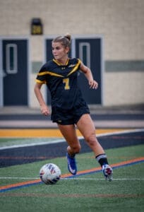 Young female soccer player in a black and yellow jersey dribbling a soccer ball on the field, focused on the game.