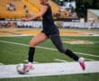 Female soccer player sprinting on the field, poised to kick the ball during a match, with spectators in the background.