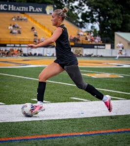 Female soccer player sprinting on the field, poised to kick the ball during a match, with spectators in the background.