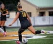 Female soccer player in action, dribbling a ball on the field during a match, wearing a black jersey and shorts.