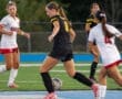 High school soccer match in progress with players in action, focusing on a girl in a black uniform dribbling the ball.