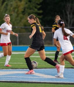 High school soccer match in progress with players in action, focusing on a girl in a black uniform dribbling the ball.