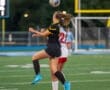 Two female soccer players jumped for a ball during a match on a field. One wears a black uniform, the other in white.