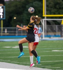 Two female soccer players jumped for a ball during a match on a field. One wears a black uniform, the other in white.