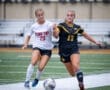 Two young female soccer players compete for the ball on a turf field during a match. Excitement and energy are evident.