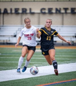 Two young female soccer players compete for the ball on a turf field during a match. Excitement and energy are evident.