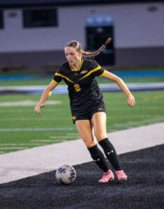 Female soccer player in a black jersey dribbling a ball on a field during a match, showcasing athletic skill and focus.
