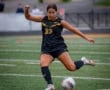 Young female soccer player in black uniform kicks a ball on a field, demonstrating athleticism and focus during a game.