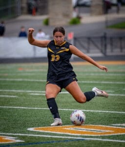 Young female soccer player in black uniform kicks a ball on a field, demonstrating athleticism and focus during a game.