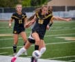 Two young female soccer players in black jerseys compete for the ball on a green field during a match.