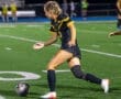 Player in black soccer uniform dribbles a ball during a night game on the field, showcasing athletic skill and focus.