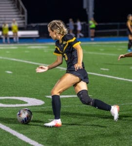 Player in black soccer uniform dribbles a ball during a night game on the field, showcasing athletic skill and focus.