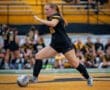 A female soccer player in a black jersey kicks a ball on a field with yellow bleachers in the background.