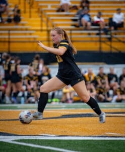 A female soccer player in a black jersey kicks a ball on a field with yellow bleachers in the background.