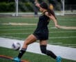 A soccer player in black uniform kicks a ball on the field during a game, showcasing athletic movement and skill.