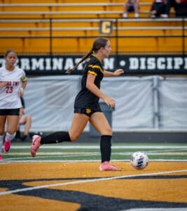Player in black jersey dribbles soccer ball on field, with teammate in white jersey nearby. Game action captured clearly.