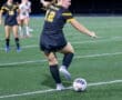High school soccer player in black uniform prepares to kick the ball during a night game on a turf field.