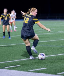 High school soccer player in black uniform prepares to kick the ball during a night game on a turf field.