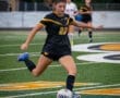 A female soccer player in black and yellow uniform dribbles a ball on the field during a game.
