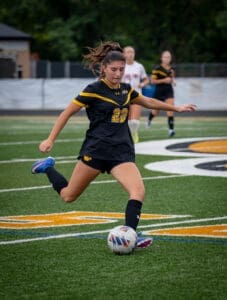 A female soccer player in black and yellow uniform dribbles a ball on the field during a game.