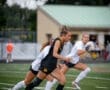Female soccer player in a black uniform sprints with a ball, while teammates compete in a dynamic match on the field.