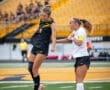 Two female soccer players compete for a header during a match, with a vibrant crowd in the background.