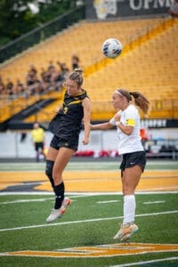 Two female soccer players compete for a header during a match, with a vibrant crowd in the background.
