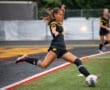 Girl in a black soccer uniform skillfully executing a kick on a grassy field during a rainy game.