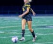 Young female soccer player in a black and yellow uniform dribbling a soccer ball on a field under bright stadium lights.