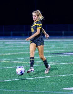 Young female soccer player in a black and yellow uniform dribbling a soccer ball on a field under bright stadium lights.