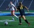 Two female soccer players compete on the field at night, with one in black and yellow, the other in white and red.