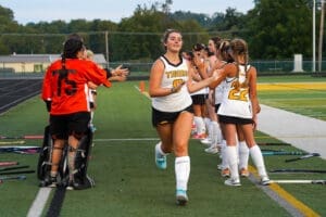 Field hockey players celebrate at the sideline as a teammate jogs through a high-five line after a successful play.