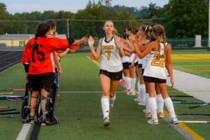 Field hockey players celebrate and cheer as a teammate in number 7 jersey is welcomed off the field after a match.
