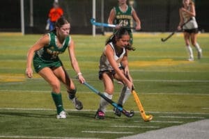 Two young female athletes compete in a high school field hockey match, showcasing action and teamwork on the field.