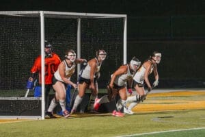 Girls' field hockey team poised for action in a match, with players and goalie preparing near the goalpost at night.