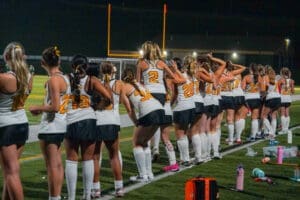 A field hockey team lines up on the sidelines, cheering under stadium lights during an evening match.