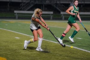 High school field hockey players in action, one striking the ball while the other advances in a night game.