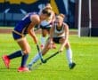 Two field hockey players compete for the ball on a sunny day, showcasing teamwork and athleticism on the turf.