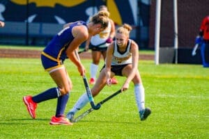 Two field hockey players compete for the ball on a sunny day, showcasing teamwork and athleticism on the turf.