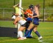 Field hockey players in action; one player deflects the ball, while a goalie prepares to block a shot.