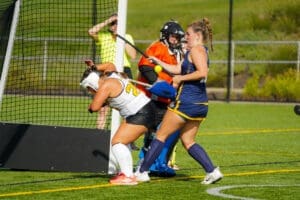 Field hockey players in action; one player deflects the ball, while a goalie prepares to block a shot.