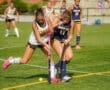 Two female field hockey players compete for the ball on a sunny day, showcasing athleticism and teamwork on the field.