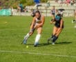 Field hockey players compete for the ball on a sunny day, with spectators watching in the background.