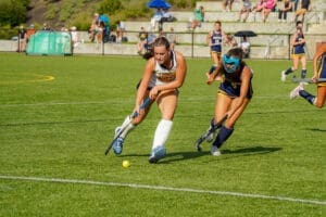 Field hockey players compete for the ball on a sunny day, with spectators watching in the background.