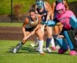 Field hockey match in action, showcasing players in motion, including a goalie in pink and a focused attacker.