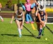 Two young female field hockey players compete for the ball on a sunny day, with a referee overseeing the match.
