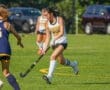 Action shot of a field hockey player in a white uniform, dribbling the ball on a sunny field during a match.