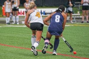 Two female field hockey players compete for the ball on a green turf field during a match.